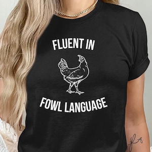 Può includere: T-shirt nera con la scritta "FLUENT IN FOWL LANGUAGE" e un'illustrazione bianca di un pollo. La maglietta è a girocollo. La persona che indossa la maglietta è visibile.