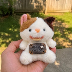 Puede incluir: Un juguete de peluche con forma de gato con pelaje blanco, marrón y canela. El gato tiene una cara sonriente, orejas rosas y una etiqueta marrón que dice "NEED COFFEE" con un icono de taza de café. El juguete se sostiene en una mano.