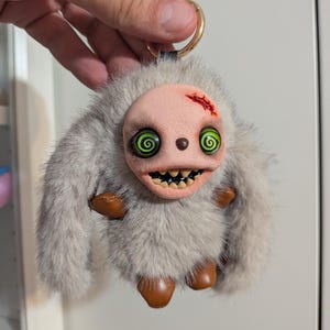 May include: A plush keyring featuring a creature with long grey faux fur, a pale pink sculpted face, and bright green spiral eyes. The figure has a small red wound detail on its forehead, sharp teeth, and brown leather-like paws and feet.