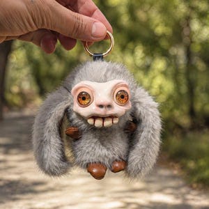 Può includere: Un portachiavi stravagante con una creatura pelosa grigia con grandi occhi arancioni e un ampio sorriso. Ha piedi e mani in similpelle marrone e un anello dorato. Lo sfondo è una scena sfocata all'aperto.