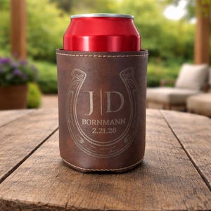 May include: A brown leather can cooler with a red can inside. The cooler features a horseshoe design with the initials "JD" and the name "BORNMANN" with the date "2.21.26" engraved on the front. The cooler is sitting on a wooden surface.