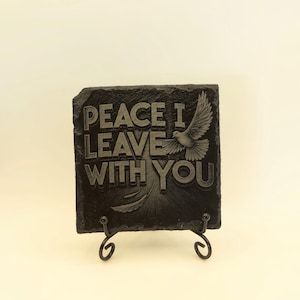 May include: A black slate plaque with the engraved text "PEACE I LEAVE WITH YOU" and a dove illustration. The plaque is displayed on a black metal stand. The dove is depicted with outstretched wings.
