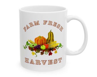 Könnte beinhalten: Weiße Keramik-Tasse mit Henkel, mit einem "Farm Fresh Harvest"-Design. Das Design umfasst einen Kürbis, eine Kürbisart, Mais, Äpfel, Trauben und Herbstblätter. Der Text ist braun.