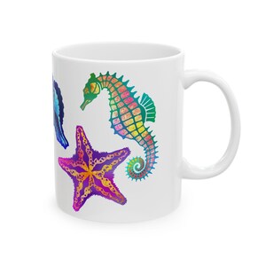 May include: White ceramic mug featuring a colourful design of a seahorse, starfish and seashell. The seahorse is green, yellow, orange and blue. The starfish is purple, yellow and orange. The seashell is blue.