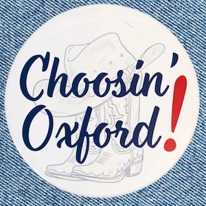 May include: A round white pin-back button featuring a light gray line drawing of a cowboy hat and a pair of cowboy boots. The text "Choosin' Oxford!" is printed in dark blue script, with the exclamation point in a bold red color.