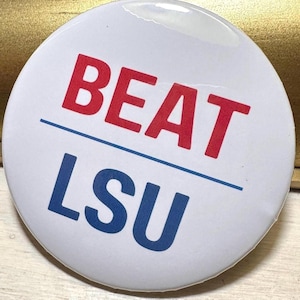 May include: A white button with the words "BEAT" in red above a blue line, and "LSU" in blue below the line. The button has a glossy finish and a simple, bold design.