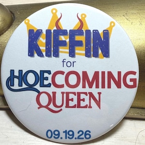 May include: A round white pin-back button featuring a yellow crown graphic and blue text that reads KIFFIN for HOECOMING QUEEN in a mix of blue and red lettering, with the date 09.19.26 printed in blue at the bottom of the circular surface.