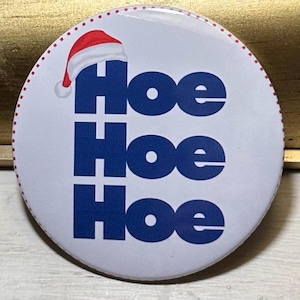 May include: A round, white button with the text "Hoe Hoe Hoe" in blue, topped with a red and white Santa hat. The button has a red dotted border and is likely a holiday-themed accessory.