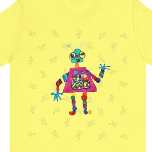 Puede incluir: Una camiseta amarilla con una ilustración de robot colorido en el centro. El robot tiene un cuerpo rosa, una cabeza verde y brazos y piernas de colores arcoíris. El fondo está cubierto de pequeños dibujos de robots negros.