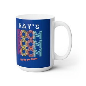 Könnte beinhalten: Weiße Keramiktasse mit blauem Hintergrund und dem Text "RAY'S ROOM" in einer Retro-Schriftart mit bunten Streifen. Der Text "The Up-per Room" befindet sich unter dem Haupttext.