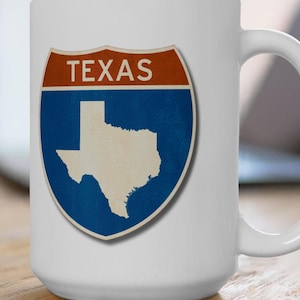 May include: White ceramic mug with a blue and red highway sign featuring the state of Texas and the word "TEXAS" in white text.