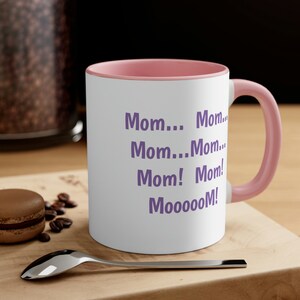 May include: A white ceramic mug with a pink rim and handle. The mug has purple text that reads "Mom... Mom... Mom... Mom! Mom! Mooooom!"