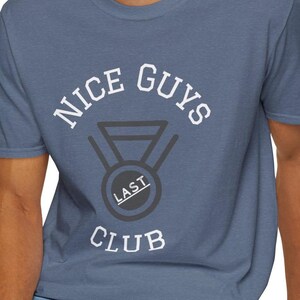 May include: A blue t-shirt with the text "NICE GUYS LAST CLUB" in white lettering. The text is arranged in a circle around a black and white graphic of a medal.