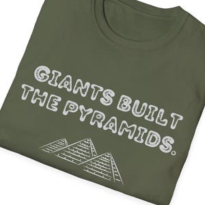 May include: Olive green t-shirt with the text "Giants Built The Pyramids" in white, along with a graphic of three pyramids. The shirt is folded, showcasing the design prominently.