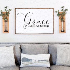 May include: A grey sofa with three cushions, one featuring a house design, in a living room with a white wall. Two wooden wall shelves with greenery and glass vases are on either side of a framed sign that reads "Grace Changes Everything".