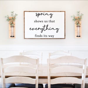 以下が含まれることがあります： 白いファームハウススタイルのサインに、黒い書体で「spring shows us that everything finds its way」と書かれています。サインは白い壁に、白いダイニングテーブルの上、白い椅子6脚の上にかけられています。サインの両側には、緑色の植物が入った小さな木製の壁掛けプランターが2つあります。