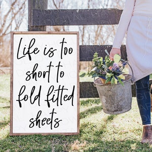 Peut inclure: Une pancarte en bois blanc avec une calligraphie noire qui dit "La vie est trop courte pour plier des draps ajustés". La pancarte est appuyée contre une clôture en bois. Une personne tient un seau de fleurs à l'arrière-plan.