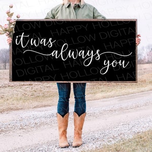May include: A black wooden sign with the text "it was always you" written in white cursive font. The sign is being held by a person wearing blue jeans and brown cowboy boots.