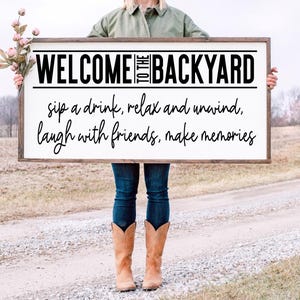 Op de afbeelding: Een wit houten bord met bruine rand heeft de tekst "WELCOME TO THE BACKYARD sip a drink, relax and unwind, laugh with friends, make memories".