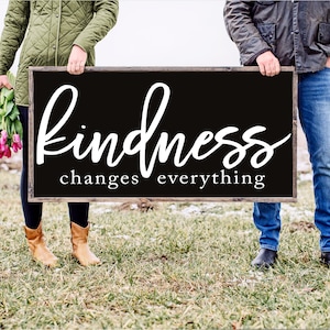May include: Two people holding a black wooden sign with white text that reads "kindness changes everything".