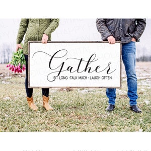 May include: A white wooden sign with black text that reads "Gather Sit Long Talk Much Laugh Often". Two people are holding the sign in front of a field.