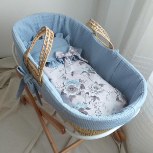 Op de afbeelding: Een blauwe en witte rieten wieg met een bloemenprint en een blauwe wafelstructuur. De wieg staat op een houten standaard met wielen.