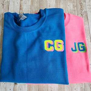 May include: Two sweatshirts, one blue and one pink, with embroidered letters in a 3D style. The blue sweatshirt has "CG" embroidered on it, and the pink sweatshirt has "JG" embroidered on it.