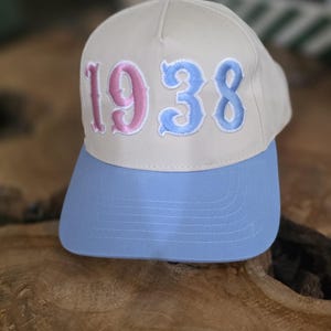 May include: A baseball cap with a light tan crown and a light blue brim. The embroidered text "1938" is in pink and blue. The hat is displayed on a wooden surface.