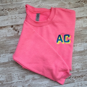 May include: A pink sweatshirt with the letters "AC" embroidered in yellow and blue.