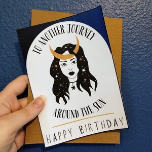 May include: A white card with a black and gold illustration of a woman with a crescent moon on her head and stars in her hair. The text on the card reads "To another journey around the sun Happy Birthday".