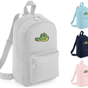 May include: A grey, blue, pink, and navy blue backpack with a cartoon crocodile face embroidered on the front. The backpack has a zipped front pocket and adjustable straps.