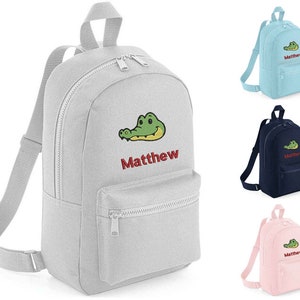 May include: A grey backpack with a green crocodile and the name Matthew embroidered on the front. The backpack has a zipped pocket on the front and two adjustable straps.