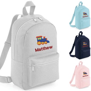 May include: A grey canvas backpack with a blue and red train embroidered on the front. The backpack has a zipped pocket on the front and two adjustable straps. The name "Matthew" is embroidered below the train.