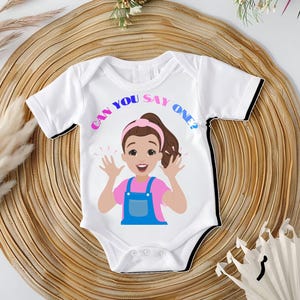 Peut inclure: Body bébé blanc avec une illustration de dessin animé d'une fille portant un t-shirt rose et une salopette bleue. Le texte "CAN YOU SAY ONE?" est en arc au-dessus de la tête de la fille.