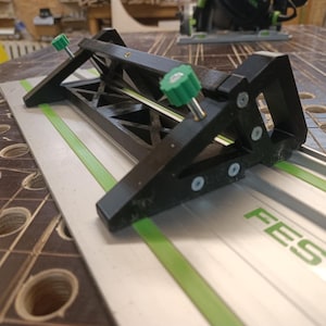 Op de afbeelding: Een zwarte en zilveren houtbewerkingsgeleider met groene accenten en verstelbare knoppen. De geleider is gepositioneerd op een zilveren rail met groene strepen en het woord "FESTOOL" in groen. De tool is ontworpen voor precisiesnijden.