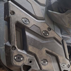 Könnte beinhalten: Nahaufnahme einer schwarzen und grauen ATV-Seitenverkleidung. Die Verkleidung hat eine strukturierte Oberfläche mit geometrischen Mustern und ist mit mehreren Schrauben und schwarzen Montageplatten befestigt. Der Boden der Verkleidung trägt den Text "4x4".