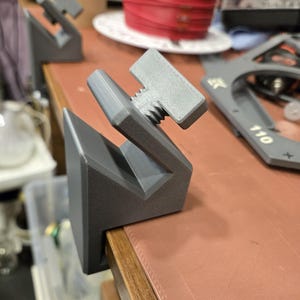 Op de afbeelding: Grijze 3D-geprinte klem met een schroefdraadmechanisme. De klem is ontworpen om aan een tafelrand te bevestigen. Het ontwerp heeft een geometrische vorm met een platte basis en schuine zijden. Gemaakt van matgrijs materiaal.