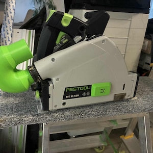 Könnte beinhalten: Eine Festool TSC 55 KEB Kreissäge mit einem grünen Staubabsauganschluss. Die Säge ist hauptsächlich grau und schwarz mit grünen Akzenten. Die Säge liegt auf einer grauen Oberfläche.
