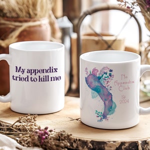 May include: Two white ceramic mugs with different designs. The mug on the left has the text "My appendix tried to kill me" in purple lettering. The mug on the right has a watercolor design of a floral bouquet with a pink and blue background. The text "No Appendix Club Est. 2024" is in pink lettering.