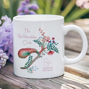 Könnte beinhalten: Weiße Keramiktasse mit einem rosafarbenen Blumendesign und dem Text "No Gallbladder Club Est. 2023".