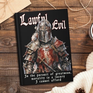 May include: A black notebook with the words "Lawful Evil" in white text. The cover features a knight in armor with a red and black design. The text below the knight reads "In the pursuit of greatness, morality is a luxury I cannot afford."