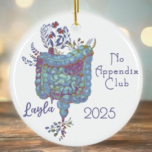 May include: White round ornament with a blue and green illustration of an intestine with flowers. The text "No Appendix Club 2025" and the name "Layla" are printed on the ornament.