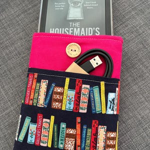 May include: A pink e-reader case with a navy blue pocket patterned with book spines. The case contains an e-reader showing "The Housemaid's" book cover and a USB cable. A wooden button with "Handmade with love" is attached.