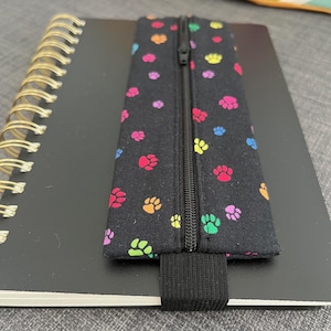 Puede incluir: Estuche negro con cremallera, adornado con coloridas huellas de patas. El estuche está sujeto a un cuaderno negro con espiral con una banda elástica negra. Las huellas de patas son en tonos rojo, naranja, amarillo, verde, azul y rosa.
