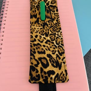 Puede incluir: Un porta bolígrafos de tela con estampado de leopardo con una banda elástica negra. El porta bolígrafos está unido a una libreta rosa.
