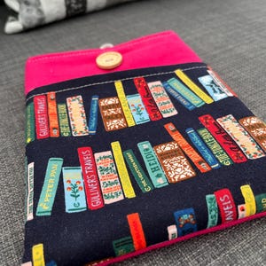 May include: A navy blue book sleeve with a pink flap and trim, featuring a colorful print of stacked books. The books have titles like "Gulliver's Travels" and "Peter Pan." A wooden button secures the flap.