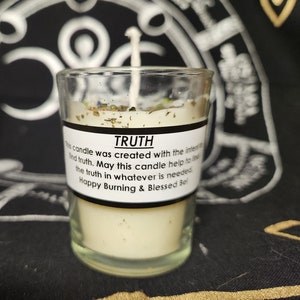 May include: A small, clear glass candle holder with a white candle inside. The candle has a white label with the word "TRUTH" and text about finding truth. The candle is decorated with dried herbs and a white wick.