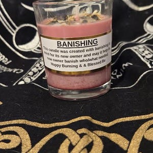 May include: A glass candle holder with a pink candle. The candle has a label that reads "BANISHING". The candle is topped with dried herbs. The candle is on a black surface with a gold pattern.