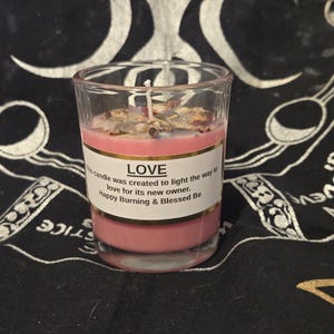 May include: A pink candle in a clear glass votive. The candle is decorated with dried herbs and flowers. A label on the glass reads "LOVE" and includes the text "candle was created to light the way to love for its new owner. Happy Burning & Blessed Be."