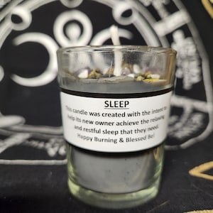 May include: A clear glass candle jar filled with gray wax and topped with small white stones and dried herbs. The label reads "SLEEP" and includes text about restful sleep. A white wick is centered in the jar.